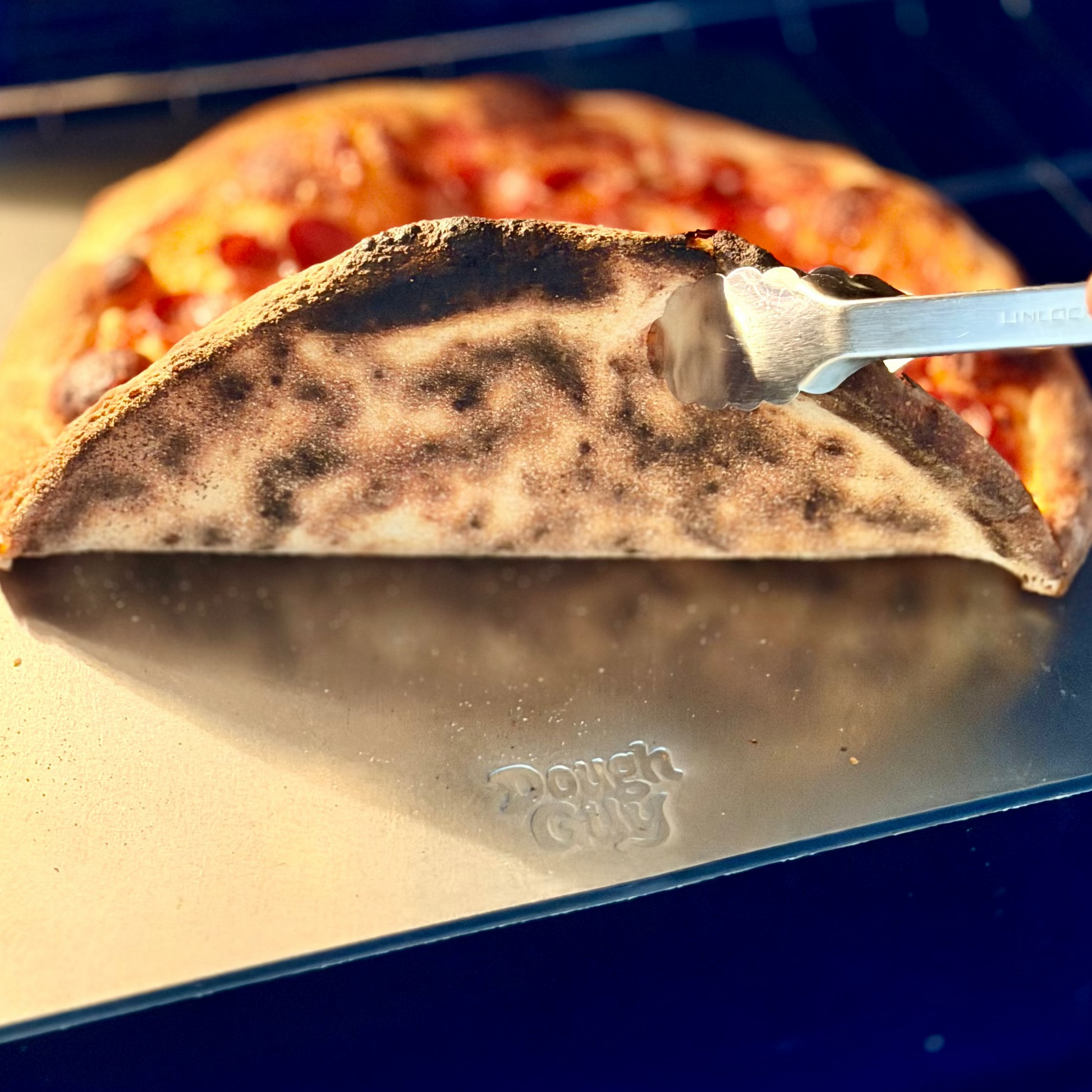 Person using tongs to remove a pizza from a pizza steel with 'Dough Guy' branding.