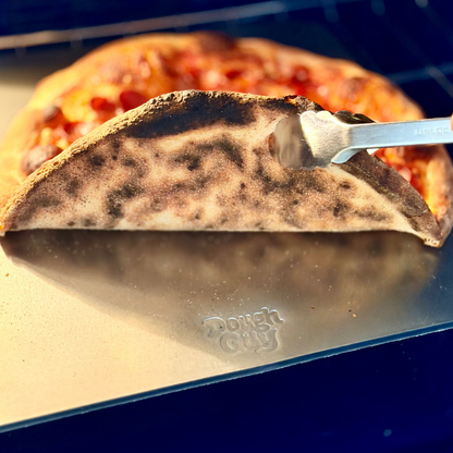 Person using tongs to remove a pizza from a pizza steel with 'Dough Guy' branding.